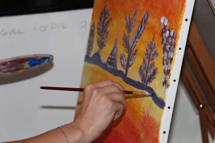 une main tenant un pinceau, en train d'ajouter de la couleur sur sa toile. La toile a un fond orangé et au premier plan on distingue des arbres et des pyramides. 